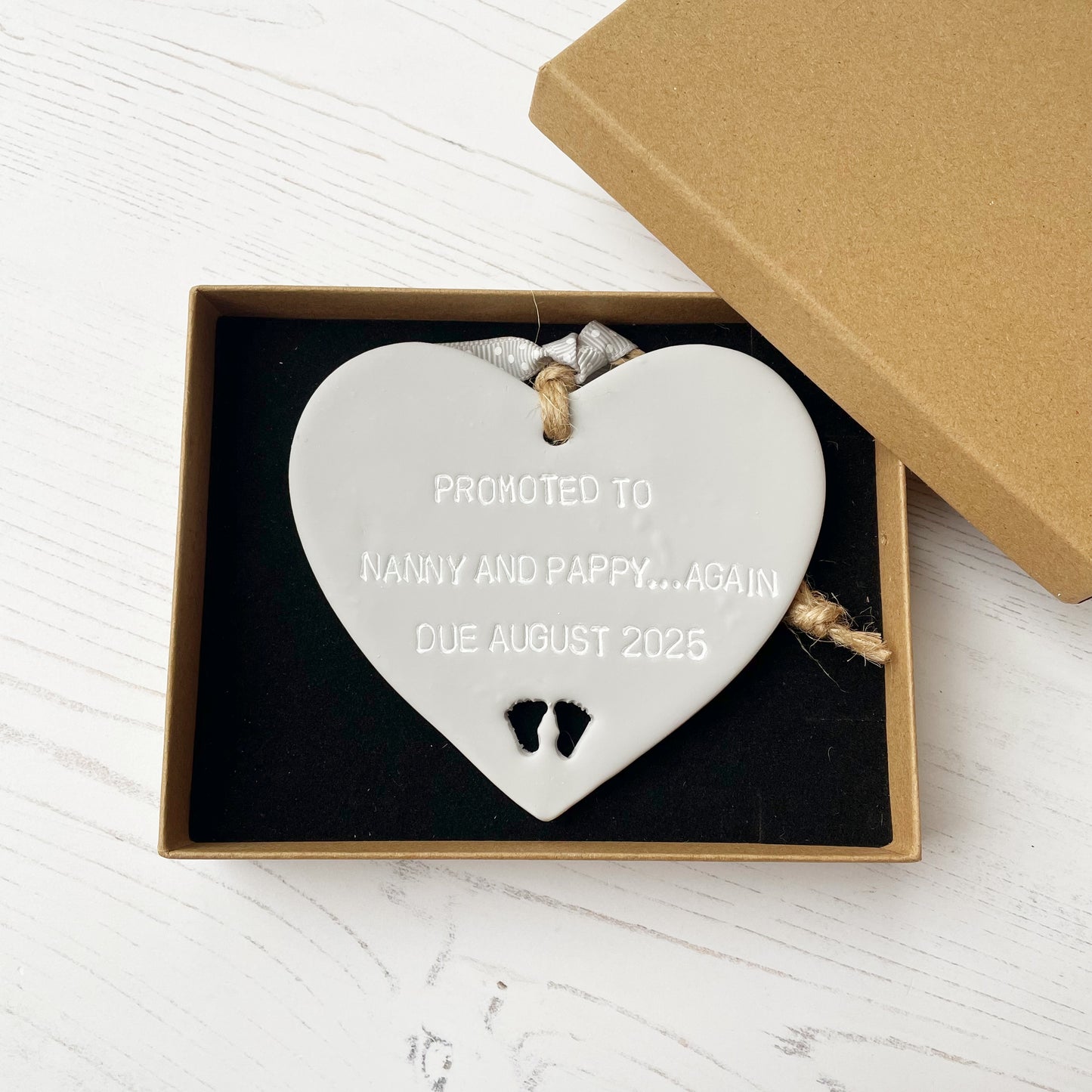 Grey clay hanging heart with baby feet cut out of the bottom and grey personalisation, the heart is personalised with PROMOTED TO NANNY AND PAPPY… AGAIN DUE AUGUST 2025