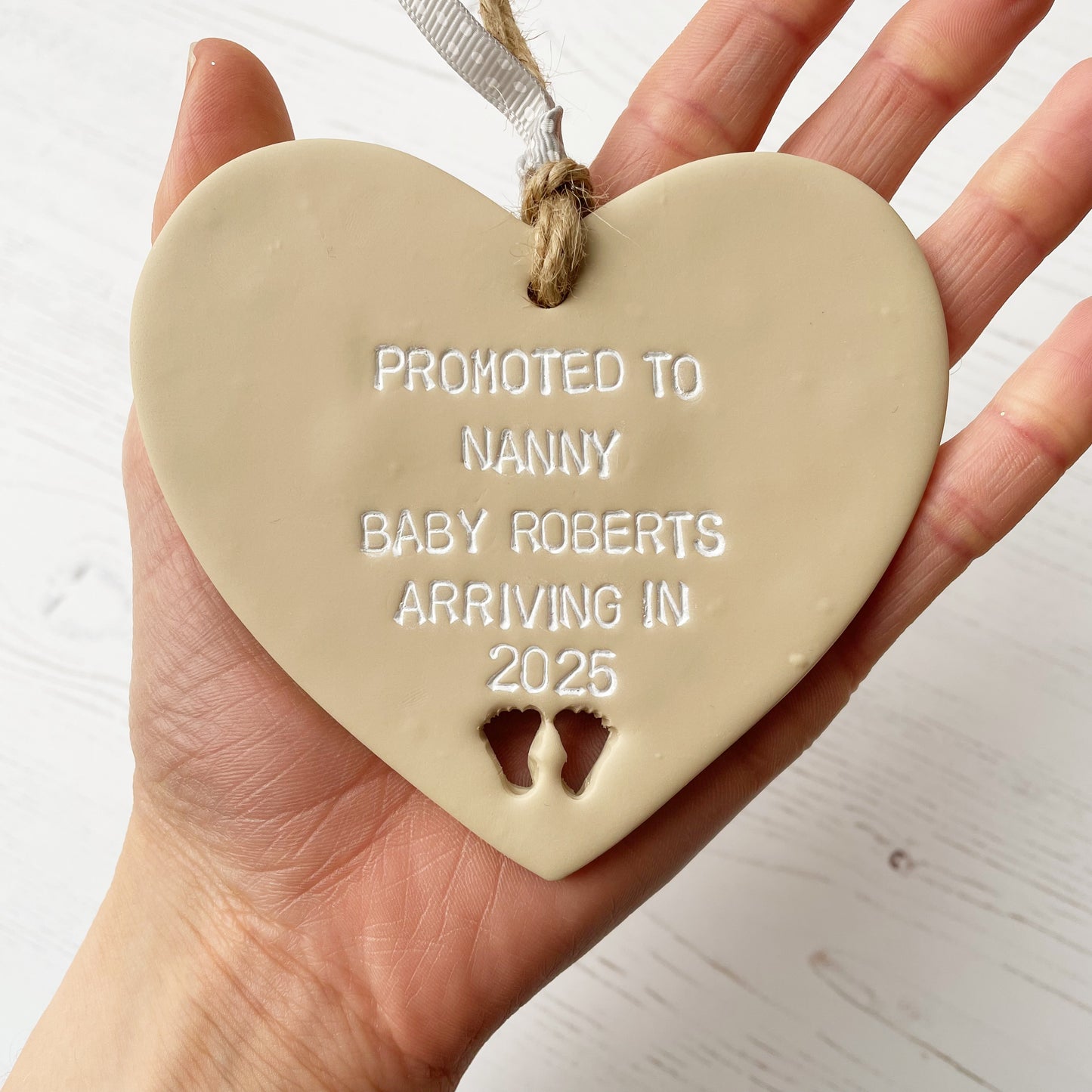 Promoted to Nanny beige clay hanging heart with baby feet cut out of the bottom and white personalisation, the heart is personalised with PROMOTED TO NANNY BABY ROBERTS ARRIVING IN 2025