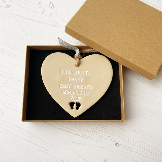 Promoted to Nanny beige clay hanging heart with baby feet cut out of the bottom and white personalisation, the heart is personalised with PROMOTED TO NANNY BABY ROBERTS ARRIVING IN 2025