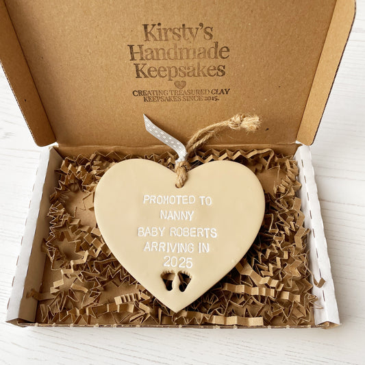 Promoted to Nanny beige clay hanging heart with baby feet cut out of the bottom and white personalisation, the heart is personalised with PROMOTED TO NANNY BABY ROBERTS ARRIVING IN 2025