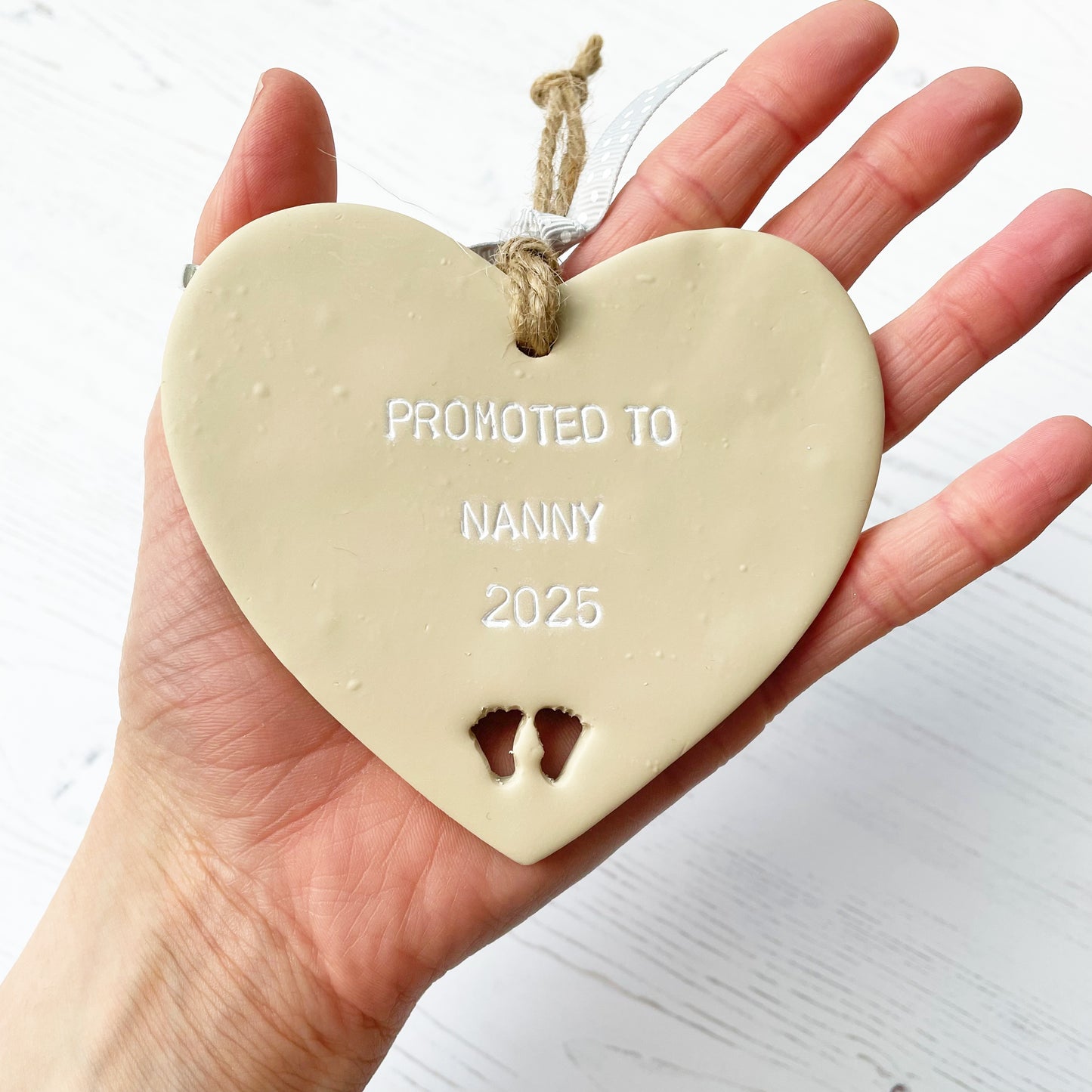 Promoted to Nanny beige clay hanging heart with baby feet cut out of the bottom and white personalisation, the heart is personalised with PROMOTED TO NANNY 2025