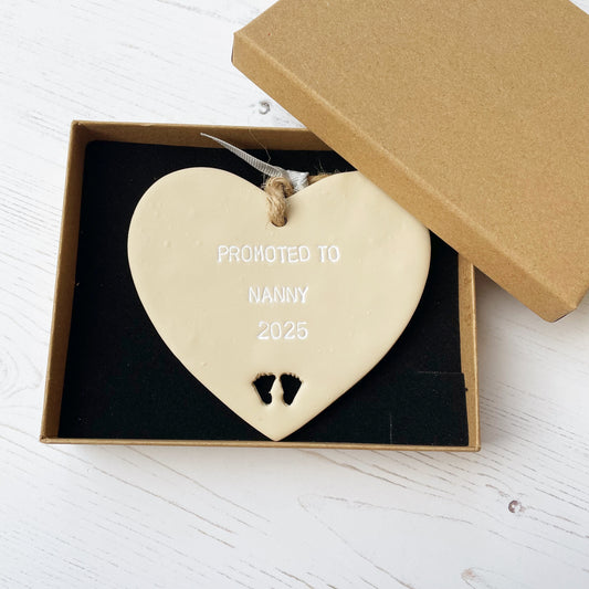 Promoted to Nanny beige clay hanging heart with baby feet cut out of the bottom and white personalisation, the heart is personalised with PROMOTED TO NANNY 2025