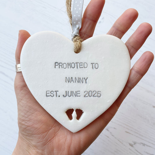 Pearlised white clay hanging heart with baby feet cut out of the bottom and grey personalisation, the heart is personalised with PROMOTED TO NANNY EST. JUNE 2025