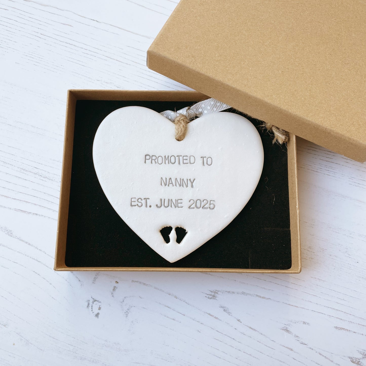 Pearlised white clay hanging heart with baby feet cut out of the bottom and grey personalisation, the heart is personalised with PROMOTED TO NANNY EST. JUNE 2025