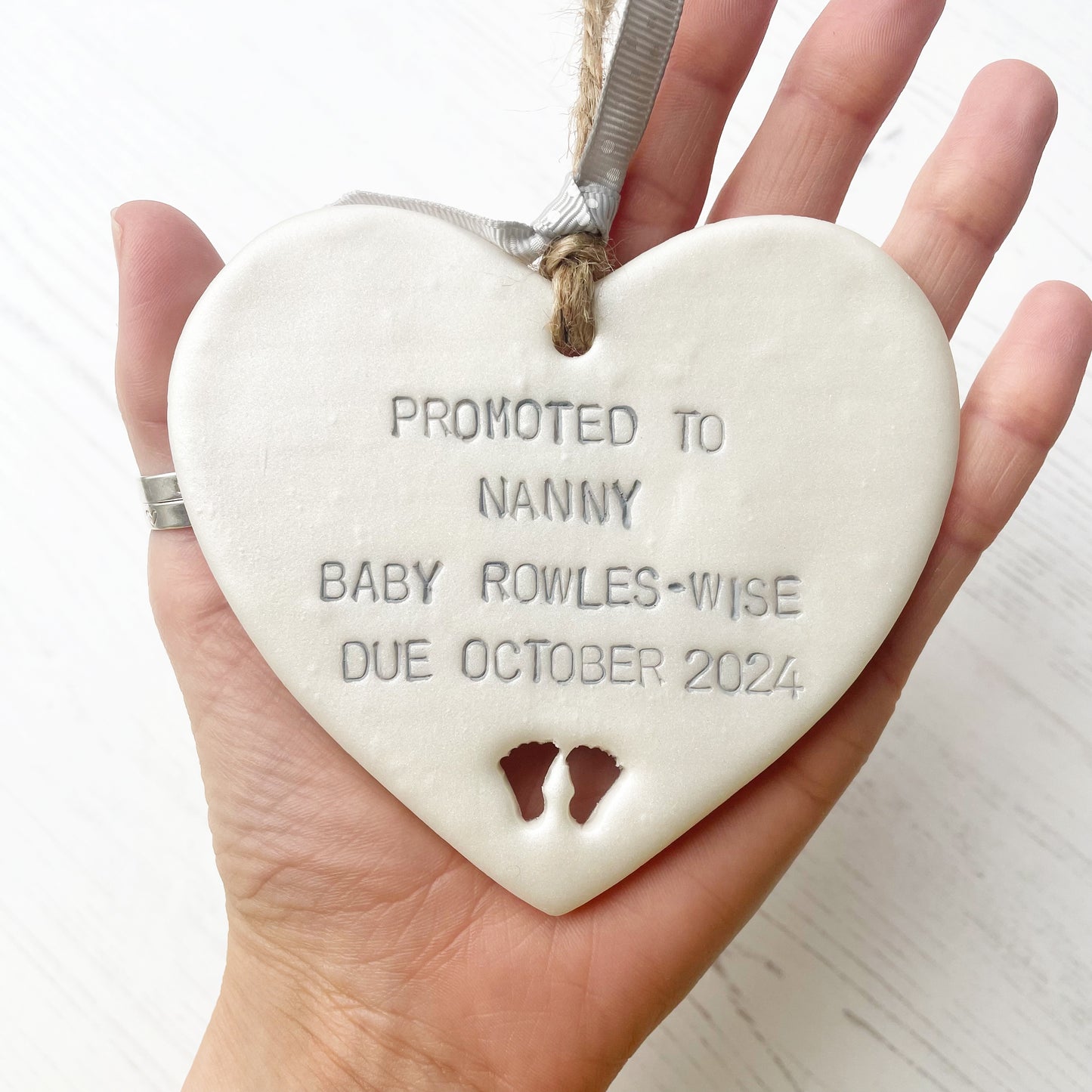 Pearlised white clay hanging heart with baby feet cut out of the bottom and grey personalisation, the heart is personalised with PROMOTED TO NANNY BABY ROWLES-WISE DUE OCTOBER 2024