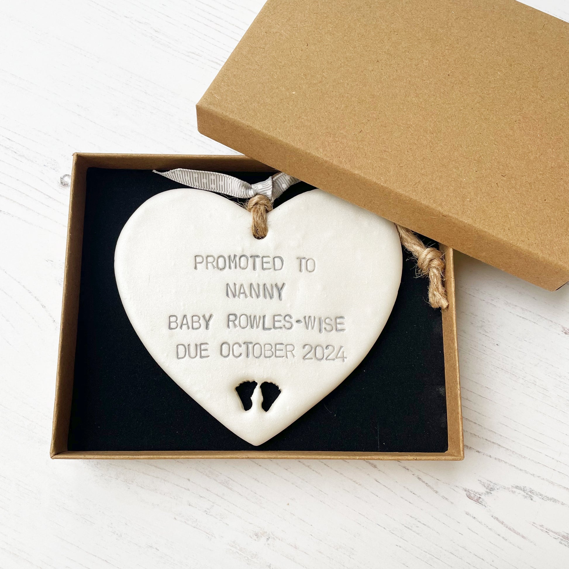 Pearlised white clay hanging heart with baby feet cut out of the bottom and grey personalisation, the heart is personalised with PROMOTED TO NANNY BABY ROWLES-WISE DUE OCTOBER 2024