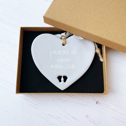 Promoted to Grandparents Grey clay hanging heart with baby feet cut out of the bottom and white personalisation, the heart is personalised with PROMOTED TO NANNY MARCH 2025