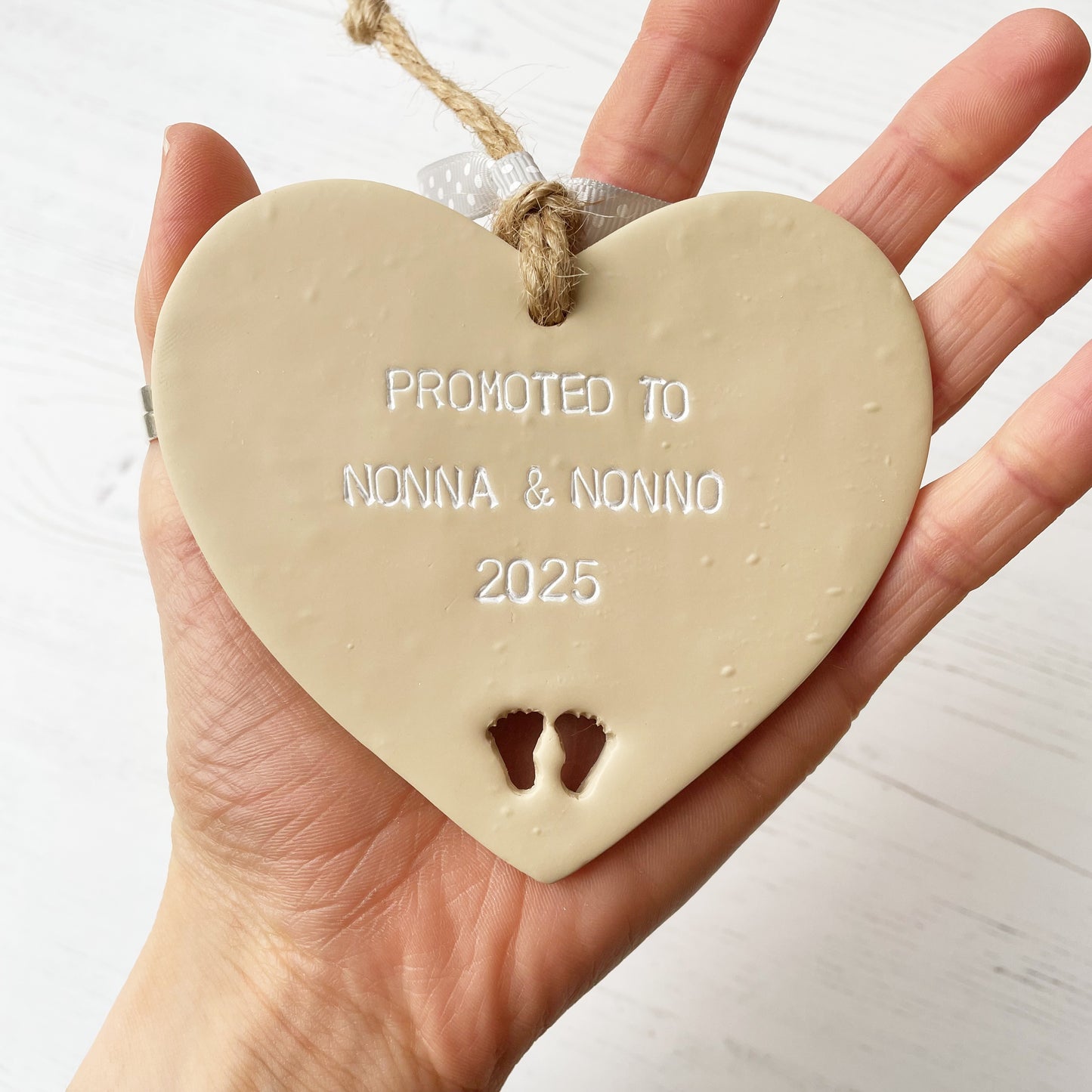 Beige clay hanging heart with baby feet cut out of the bottom and white personalisation, the heart is personalised with PROMOTED TO NONNA AND NONNO 2025