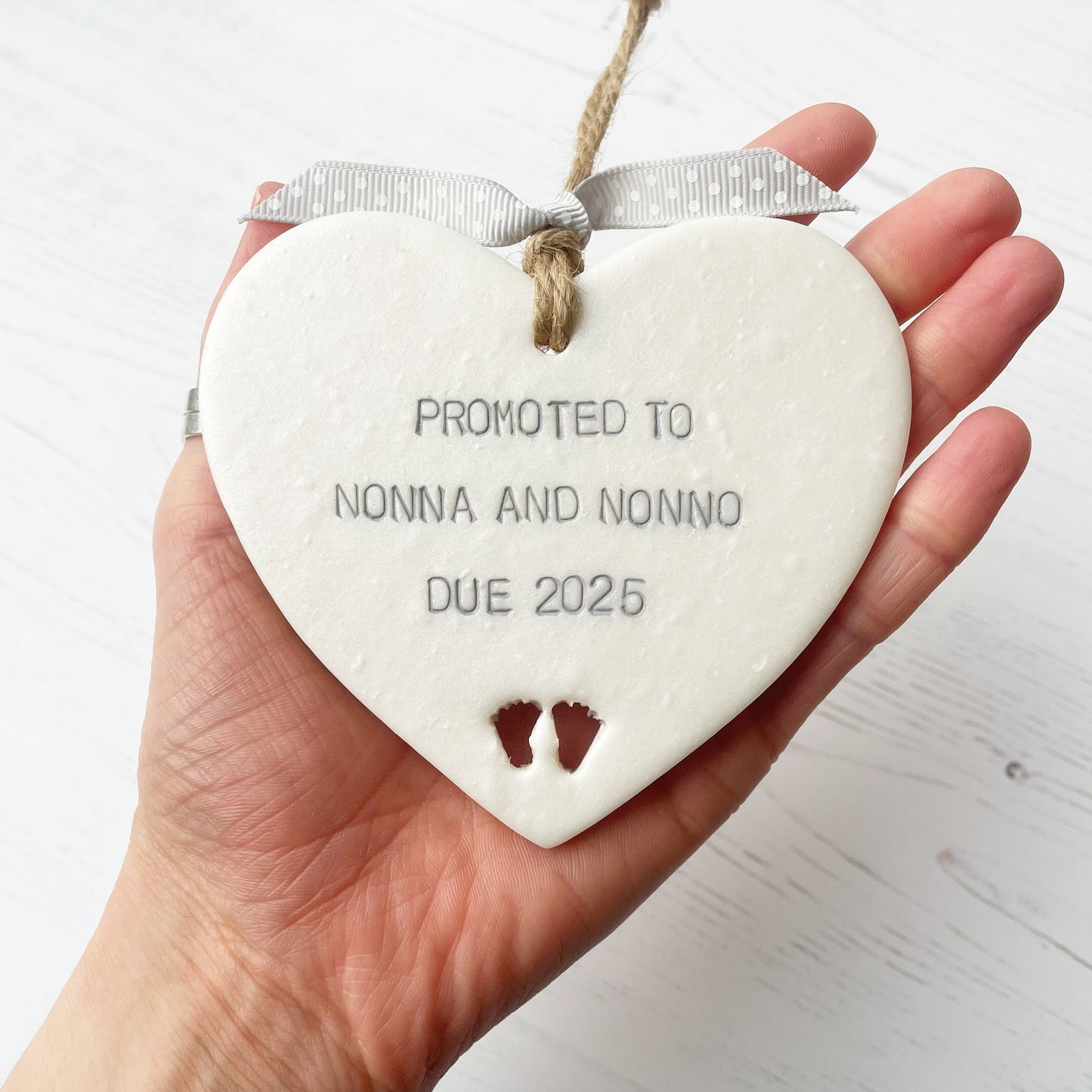 Pearlised white clay hanging heart with baby feet cut out of the bottom and grey personalisation, the heart is personalised with PROMOTED TO NONNA AND NONNO DUE 2025