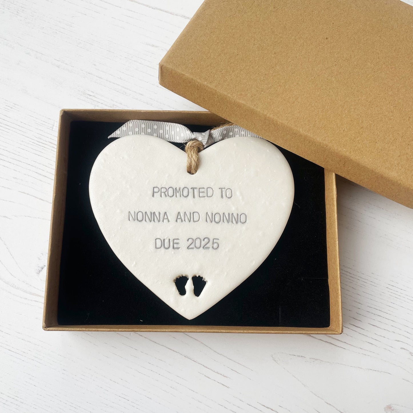 Pearlised white clay hanging heart with baby feet cut out of the bottom and grey personalisation, the heart is personalised with PROMOTED TO NONNA AND NONNO DUE 2025