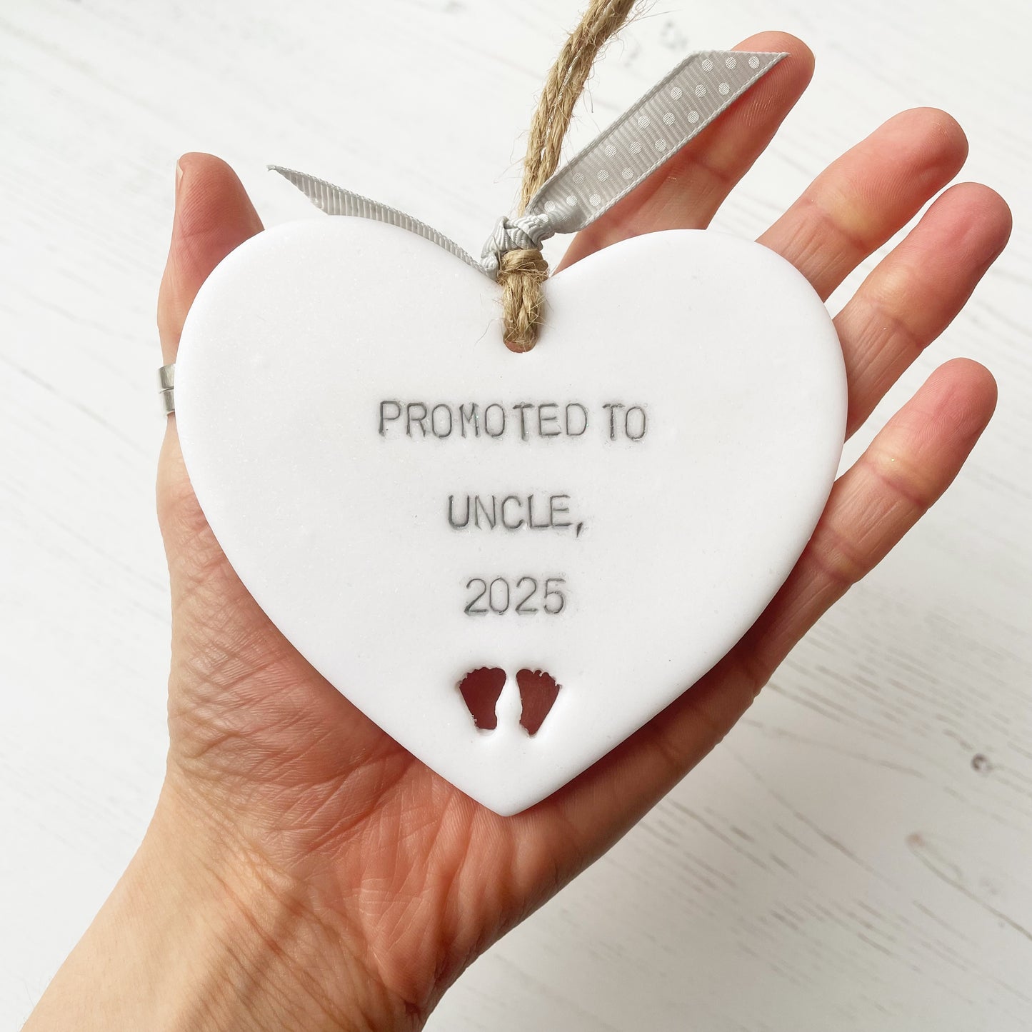 Pearlised white clay hanging heart with baby feet cut out of the bottom and grey personalisation, the heart is personalised with PROMOTED TO UNCLE, 2025