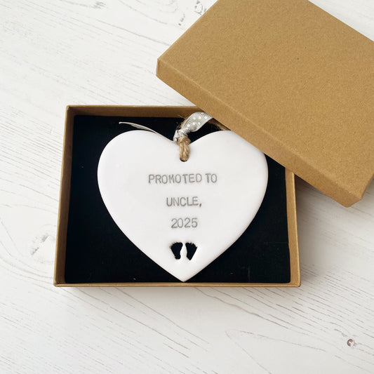 Pearlised white clay hanging heart with baby feet cut out of the bottom and grey personalisation, the heart is personalised with PROMOTED TO UNCLE, 2025