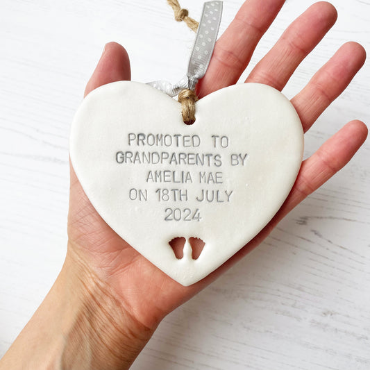 Pearlised white clay hanging heart with baby feet cut out of the bottom and grey personalisation, the heart is personalised with PROMOTED TO GRANDPARENTS BY AMELIA MAE ON 18TH JULY 2024