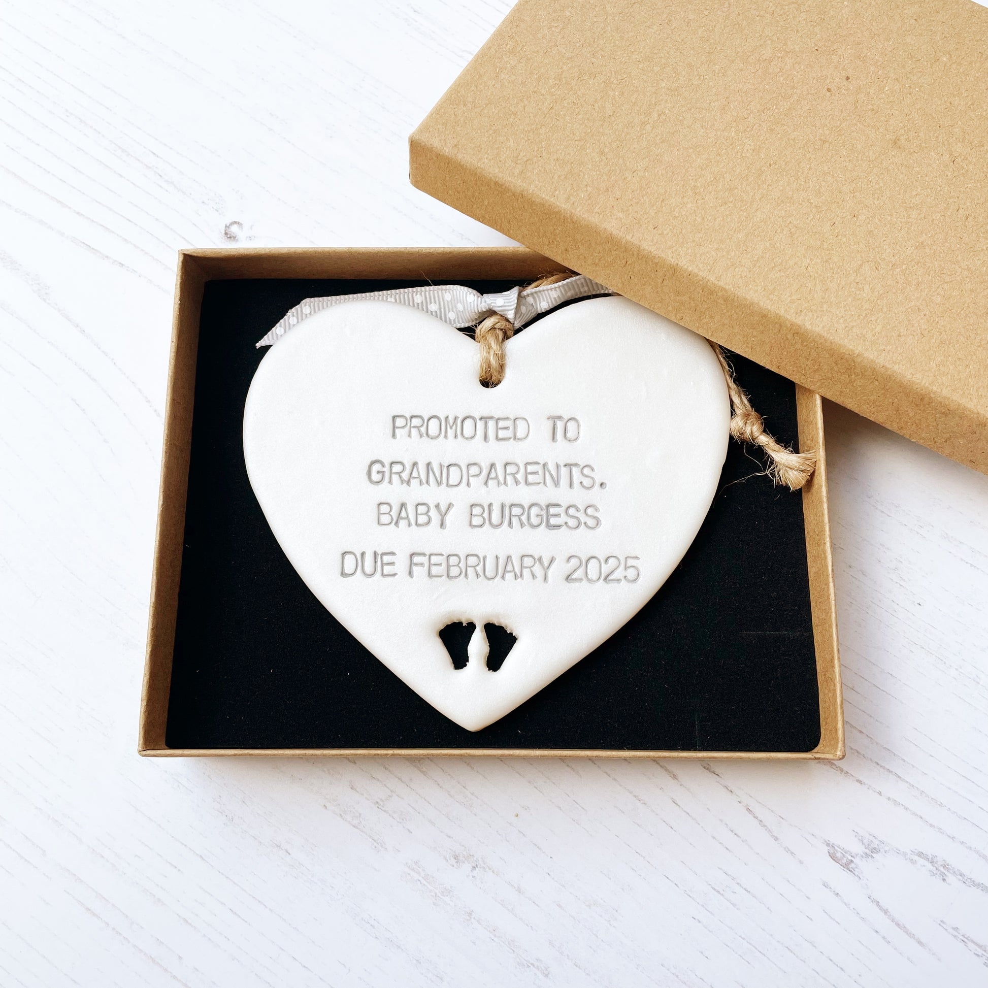 Pearlised white clay hanging heart with baby feet cut out of the bottom and grey personalisation, the heart is personalised with PROMOTED TO GRANDPARENTS. BABY BURGESS DUE FEBRUARY 2025