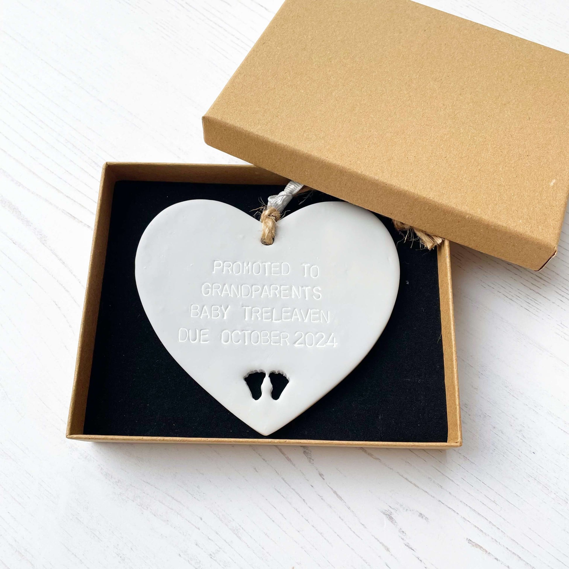 Grey clay hanging heart with baby feet cut out of the bottom and grey personalisation, the heart is personalised with PROMOTED TO GRANDPARENTS BABY TRELEAVEN DUE OCTOBER 2024