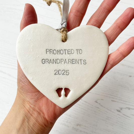 Pearlised white clay hanging heart with baby feet cut out of the bottom and grey personalisation, the heart is personalised with PROMOTED TO GRANDPARENTS 2025