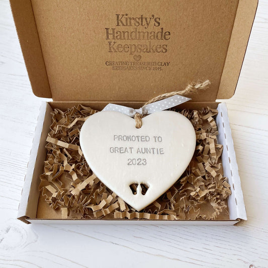 Pearlised white clay hanging heart with baby feet cut out of the bottom and grey personalisation, the heart is personalised with PROMOTED TO GREAT AUNTIE 2023