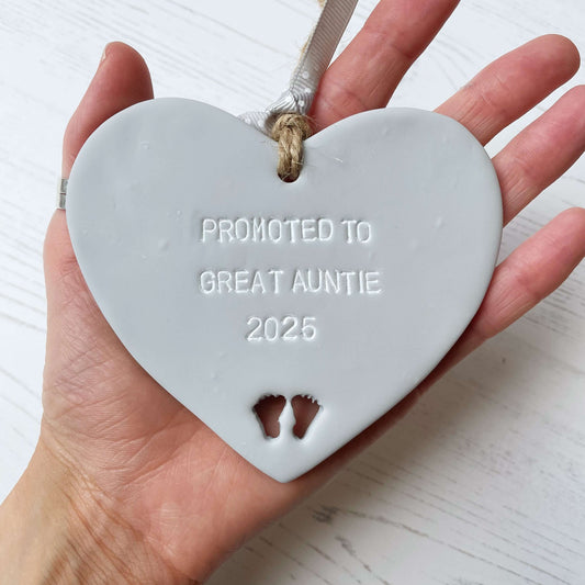 Grey clay hanging heart with baby feet cut out of the bottom and white personalisation, the heart is personalised with PROMOTED TO GREAT AUNTIE 2025