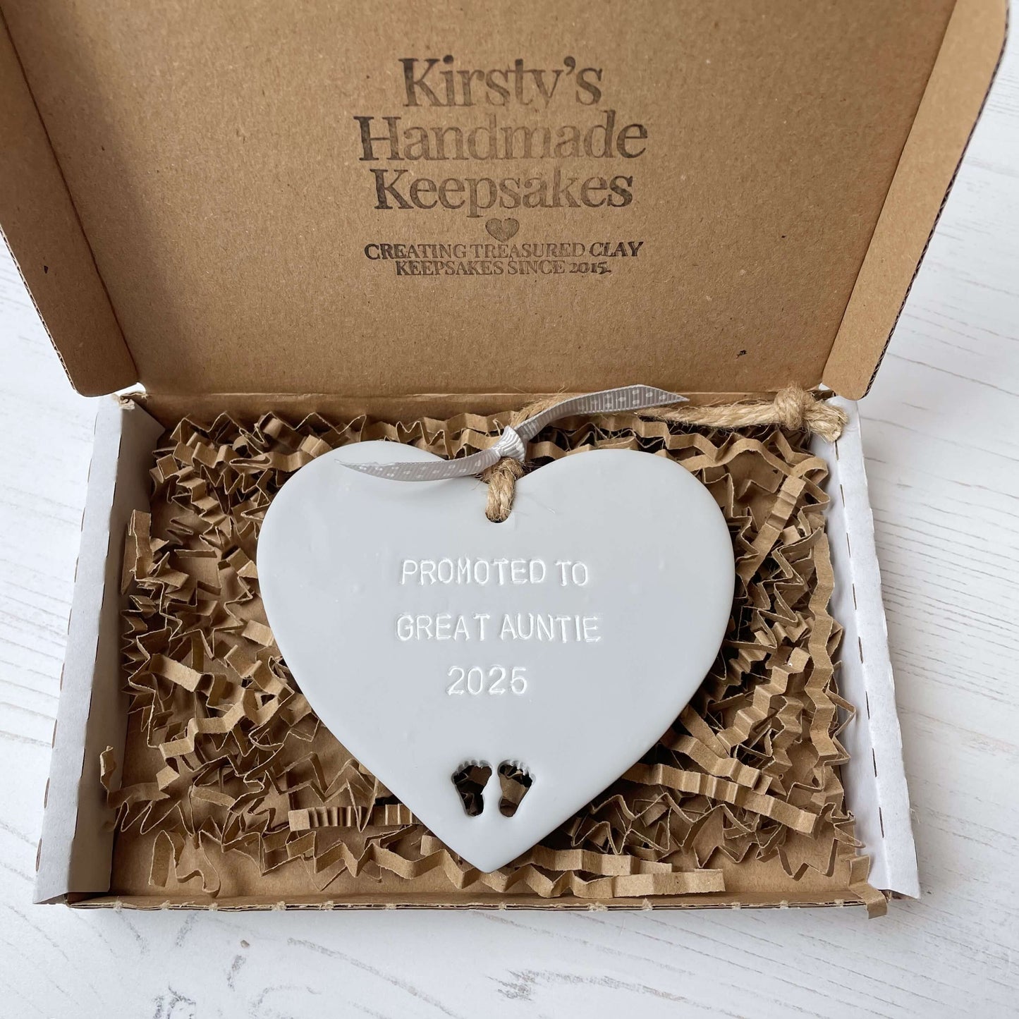 Grey clay hanging heart with baby feet cut out of the bottom and white personalisation, the heart is personalised with PROMOTED TO GREAT AUNTIE 2025