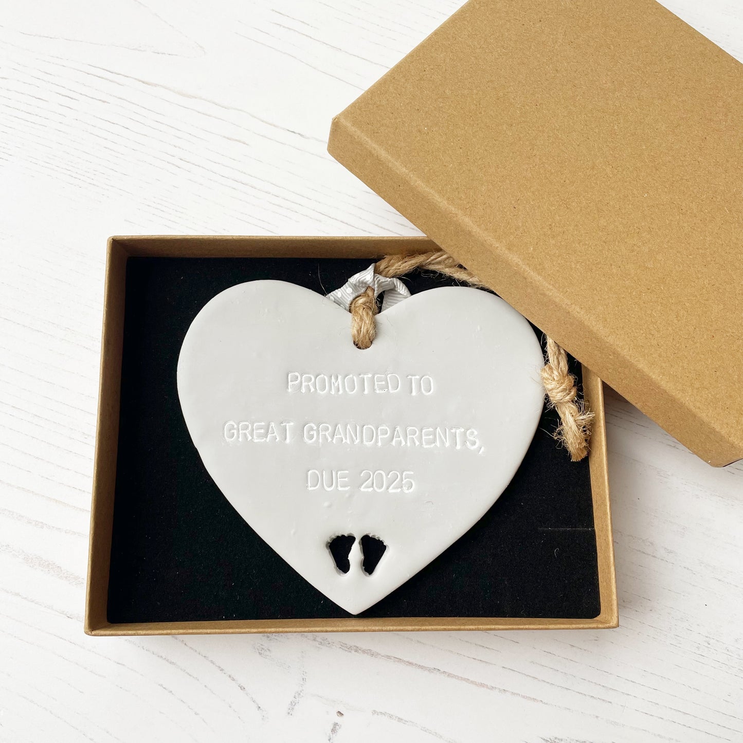 Grey clay hanging heart with baby feet cut out of the bottom and white personalisation, the heart is personalised with PROMOTED TO GREAT GRANDPARENTS DUE 2025