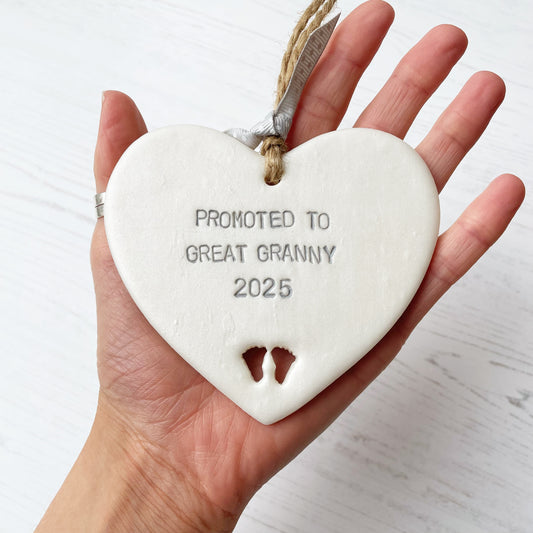 Pearlised white clay hanging heart with baby feet cut out of the bottom and grey personalisation, the heart is personalised with PROMOTED TO GREAT GRANNY 2025