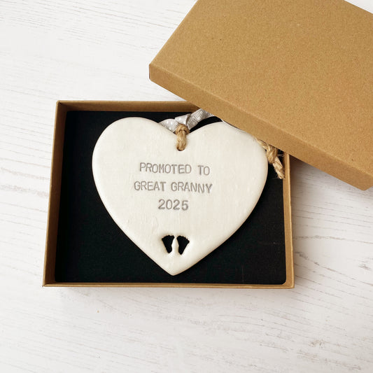 Pearlised white clay hanging heart with baby feet cut out of the bottom and grey personalisation, the heart is personalised with PROMOTED TO GREAT GRANNY 2025