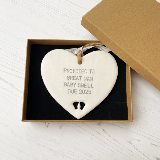 Pearlised white clay hanging heart with baby feet cut out of the bottom and grey personalisation, the heart is personalised with PROMOTED TO GREAT NAN BABY SNELL DUE 2025