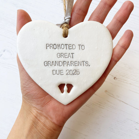 Pearlised white clay hanging heart with baby feet cut out of the bottom and grey personalisation, the heart is personalised with PROMOTED TO GREAT GRANDPARENTS. DUE 2025