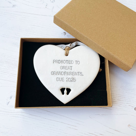 Pearlised white clay hanging heart with baby feet cut out of the bottom and grey personalisation, the heart is personalised with PROMOTED TO GREAT GRANDPARENTS. DUE 2025