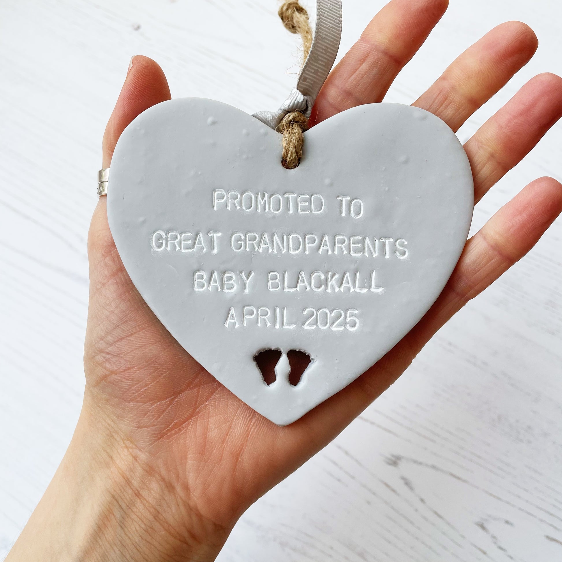 Grey clay hanging heart with baby feet cut out of the bottom and white personalisation, the heart is personalised with PROMOTED TO GREAT GRANDPARENTS BABY BLACKALL APRIL 2025