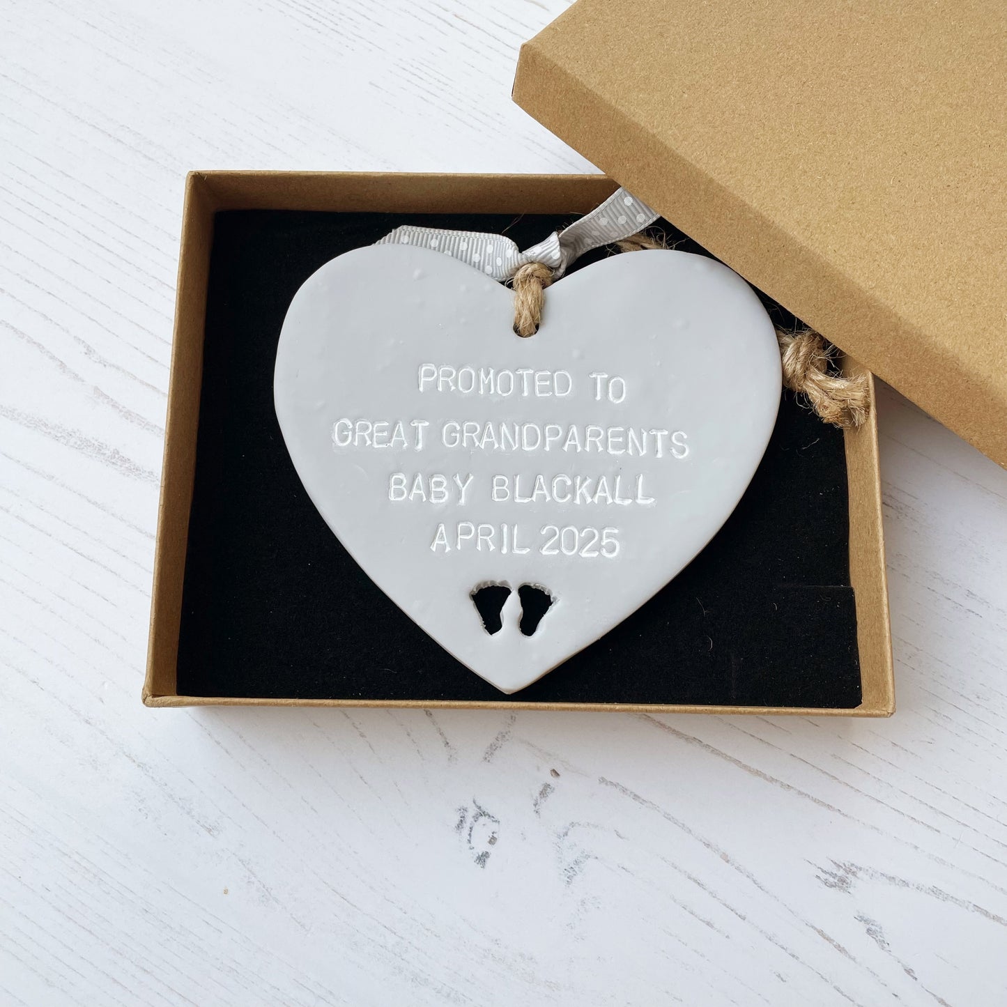 Grey clay hanging heart with baby feet cut out of the bottom and white personalisation, the heart is personalised with PROMOTED TO GREAT GRANDPARENTS BABY BLACKALL APRIL 2025