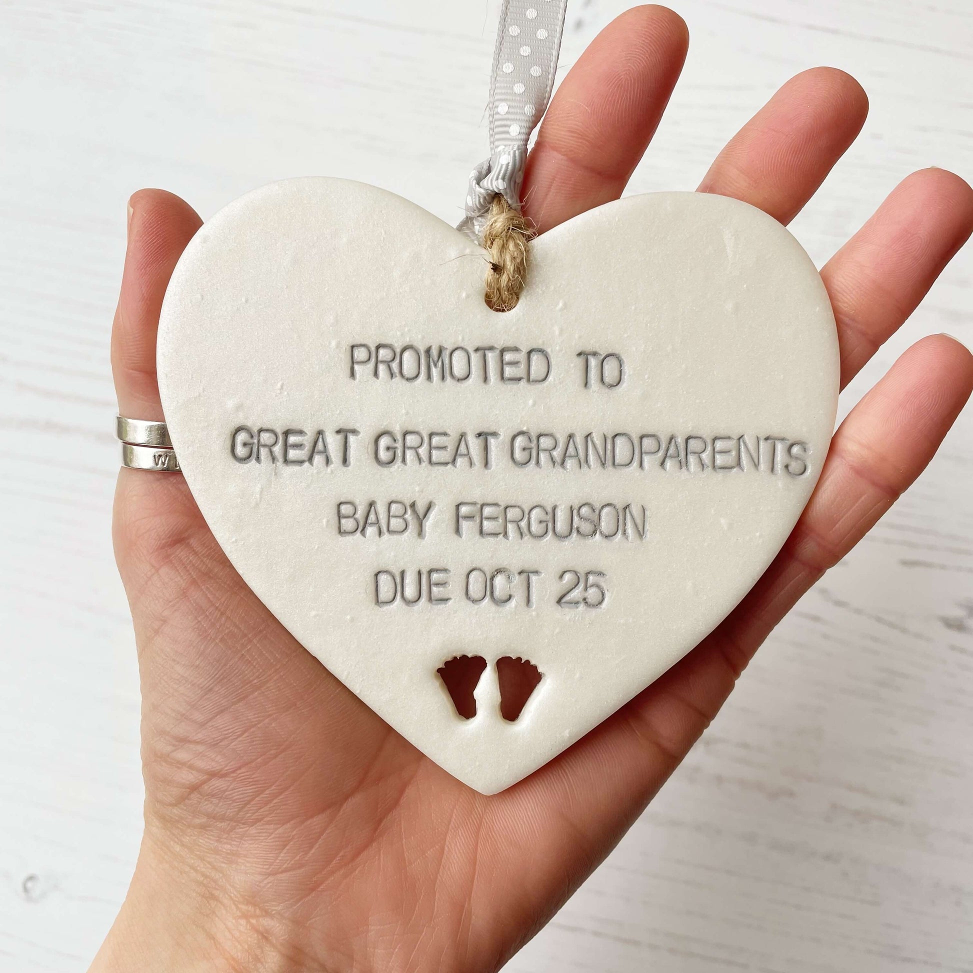 Pearlised white clay hanging heart with baby feet cut out of the bottom and grey personalisation, the heart is personalised with PROMOTED TO GREAT GREAT GRANDPARENTS BABY FERGUSON DUE OCT 2025