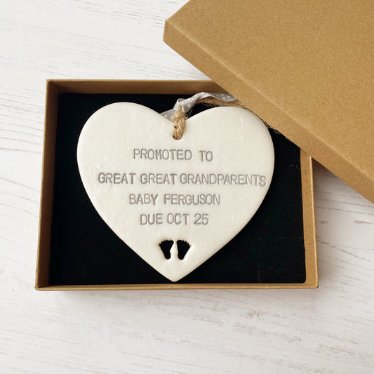 Pearlised white clay hanging heart with baby feet cut out of the bottom and grey personalisation, the heart is personalised with PROMOTED TO GREAT GREAT GRANDPARENTS BABY FERGUSON DUE OCT 2025