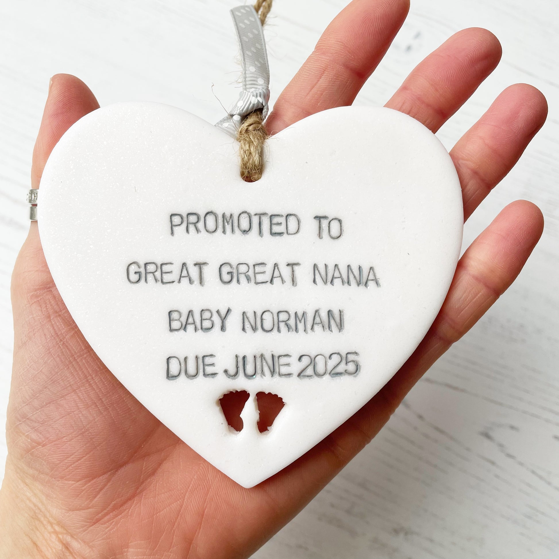 Pearlised white clay hanging heart with baby feet cut out of the bottom and grey personalisation, the heart is personalised with PROMOTED TO GREAT GREAT NANA BABY NORMAN DUE JUNE 2025