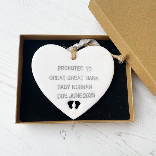 Pearlised white clay hanging heart with baby feet cut out of the bottom and grey personalisation, the heart is personalised with PROMOTED TO GREAT GREAT NANA BABY NORMAN DUE JUNE 2025