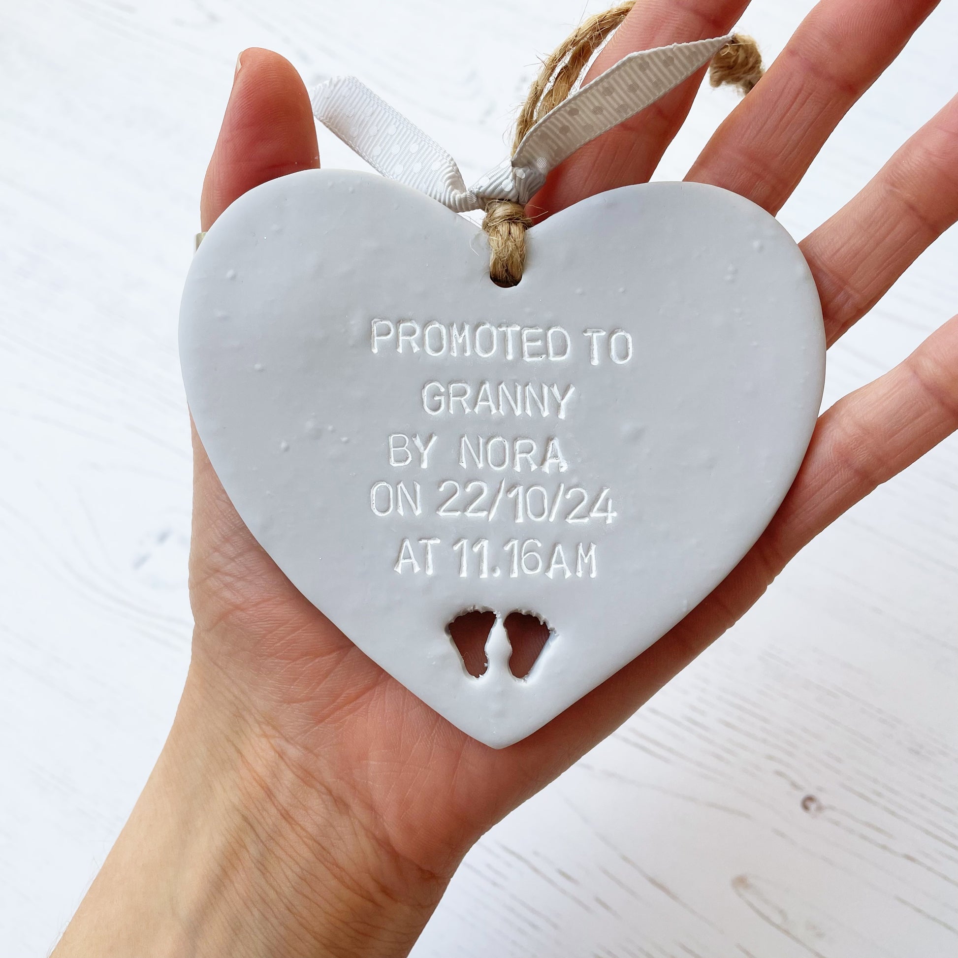 Grey clay hanging heart with baby feet cut out of the bottom and white personalisation, the heart is personalised with PROMOTED TO GRANNY BY NORA ON  22/10/24 AT 11.16 AM