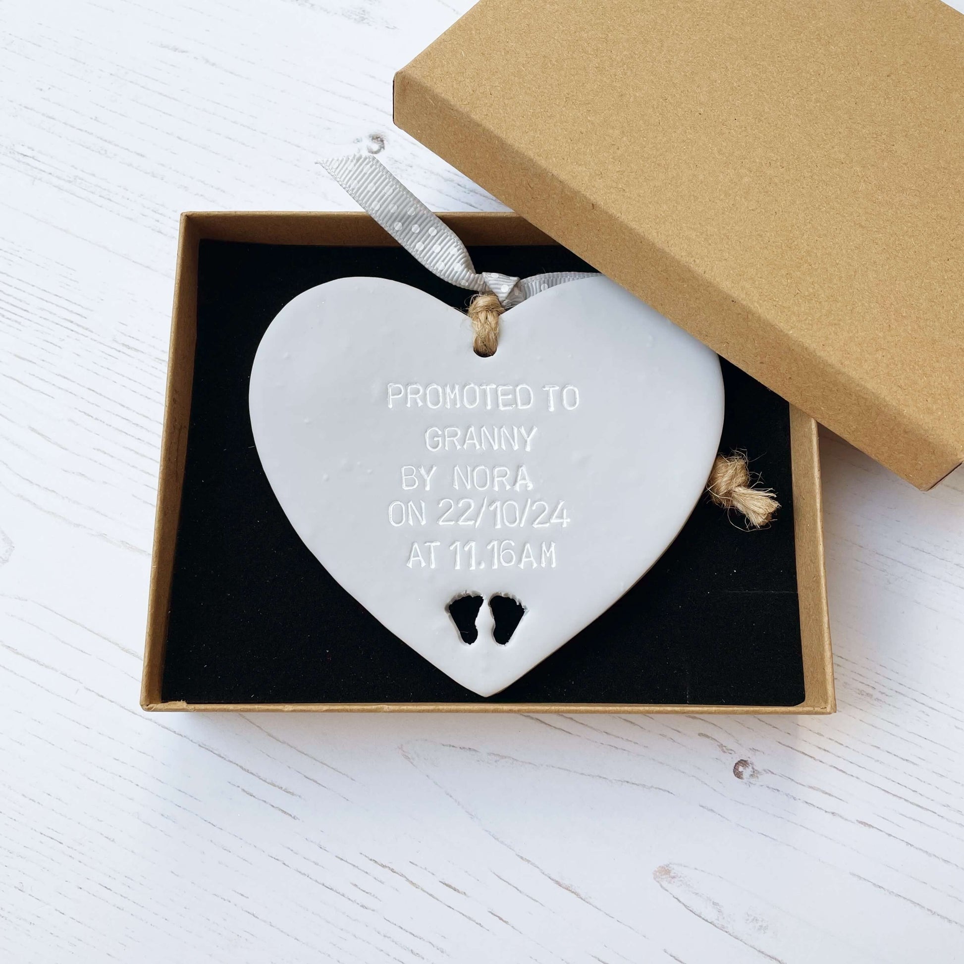 Grey clay hanging heart with baby feet cut out of the bottom and white personalisation, the heart is personalised with PROMOTED TO GRANNY BY NORA ON  22/10/24 AT 11.16 AM