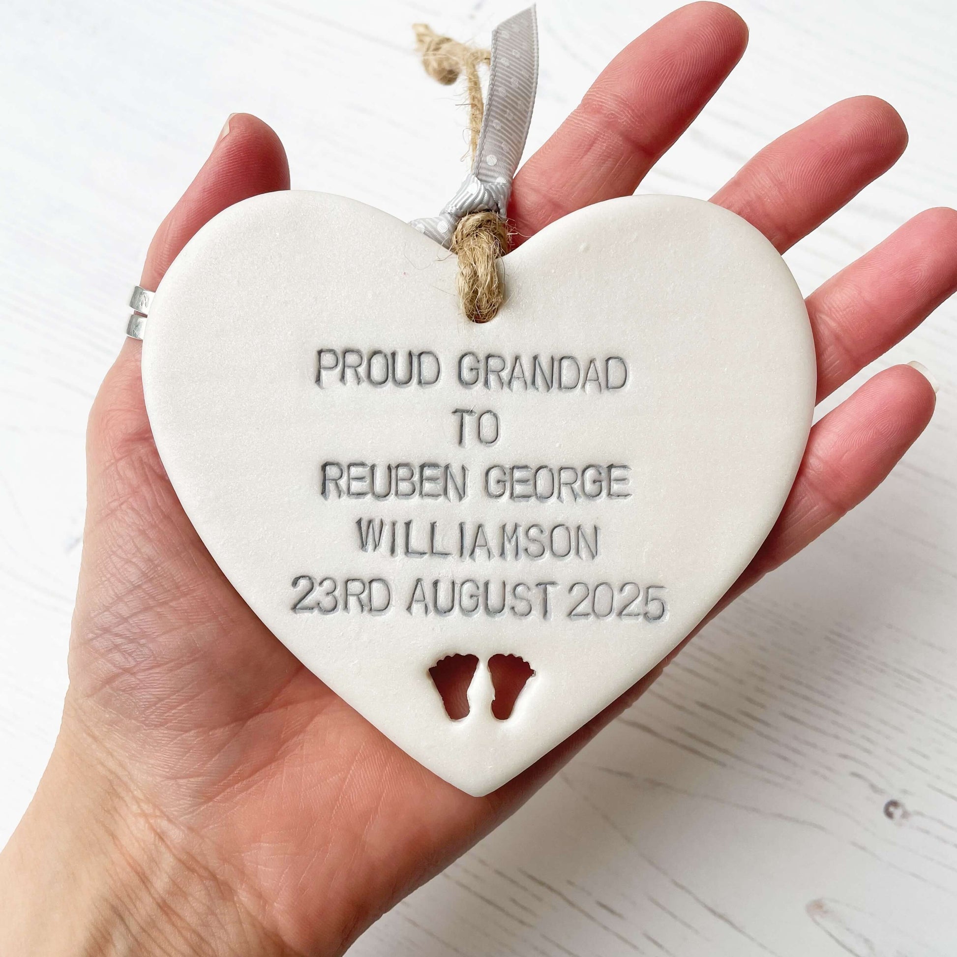Pearlised white clay hanging heart with baby feet cut out of the bottom and grey personalisation, the heart is personalised with PROUD GRANDAD TO REUBEN GEORGE WILLIAMSON 23RD AUGUST 2025