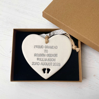 Pearlised white clay hanging heart with baby feet cut out of the bottom and grey personalisation, the heart is personalised with PROUD GRANDAD TO REUBEN GEORGE WILLIAMSON 23RD AUGUST 2025