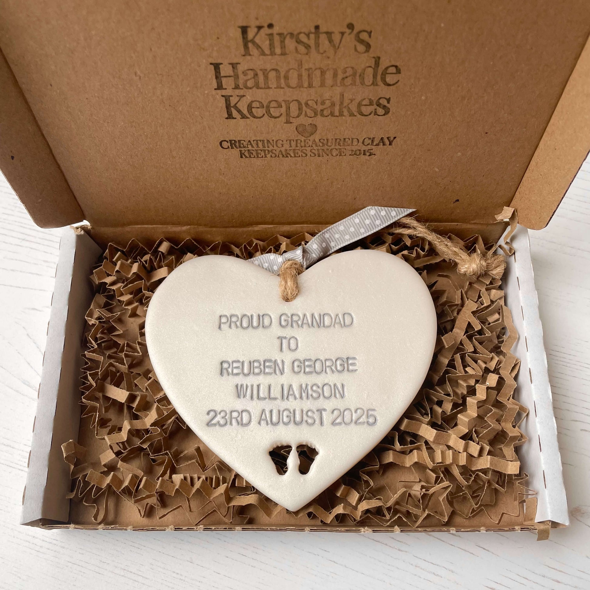 Pearlised white clay hanging heart with baby feet cut out of the bottom and grey personalisation, the heart is personalised with PROUD GRANDAD TO REUBEN GEORGE WILLIAMSON 23RD AUGUST 2025