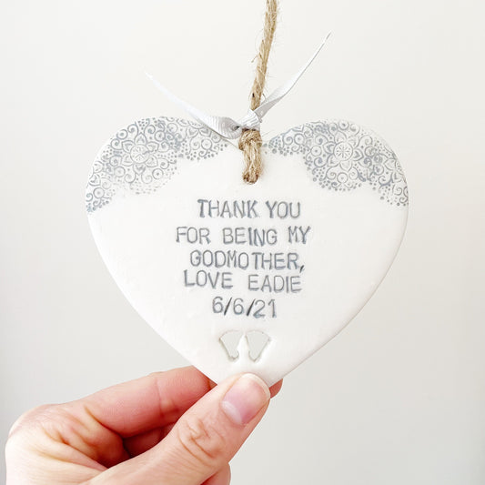 Personalised Godmother gift, pearlised white clay hanging heart with baby feet cut out of the bottom and grey lace edge at the top of the heart, the heart is personalised with THANK YOU FOR BEING MY GODMOTHER, LOVE EADIE 6/6/21