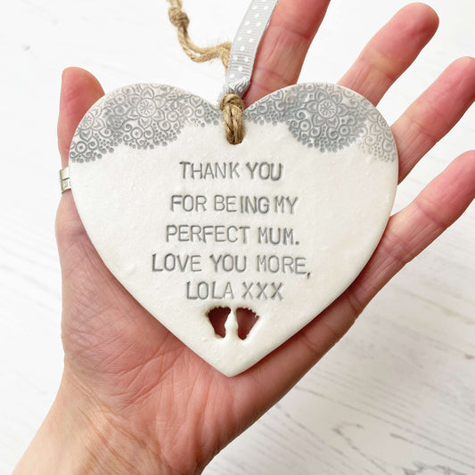 Personalised Mother’s Day gift, pearlised white clay hanging heart with baby feet cut out at the bottom of the heart and a grey lace edge at the top of the heart, the heart is personalised with THANK YOU FOR BEING MY PERFECT MUM. LOVE YOU MORE, LOLA XXX