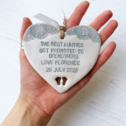 Personalised Godmother gift, pearlised white clay hanging heart with baby feet cut out of the bottom and grey lace edge at the top of the heart, the heart is personalised with THE BEST AUNTIES GET PROMOTED TO GODMOTHERS LOVE FLORENCE 26 JULY 2026