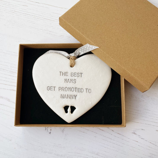 Pearlised white clay hanging heart with baby feet cut out of the bottom and grey personalisation, the heart is personalised with THE BEST MAMS GET PROMOTED TO NANNY