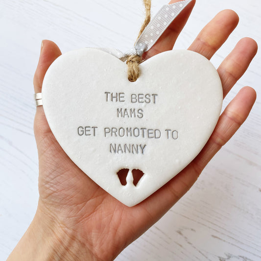 Pearlised white clay hanging heart with baby feet cut out of the bottom and grey personalisation, the heart is personalised with THE BEST MAMS GET PROMOTED TO NANNY