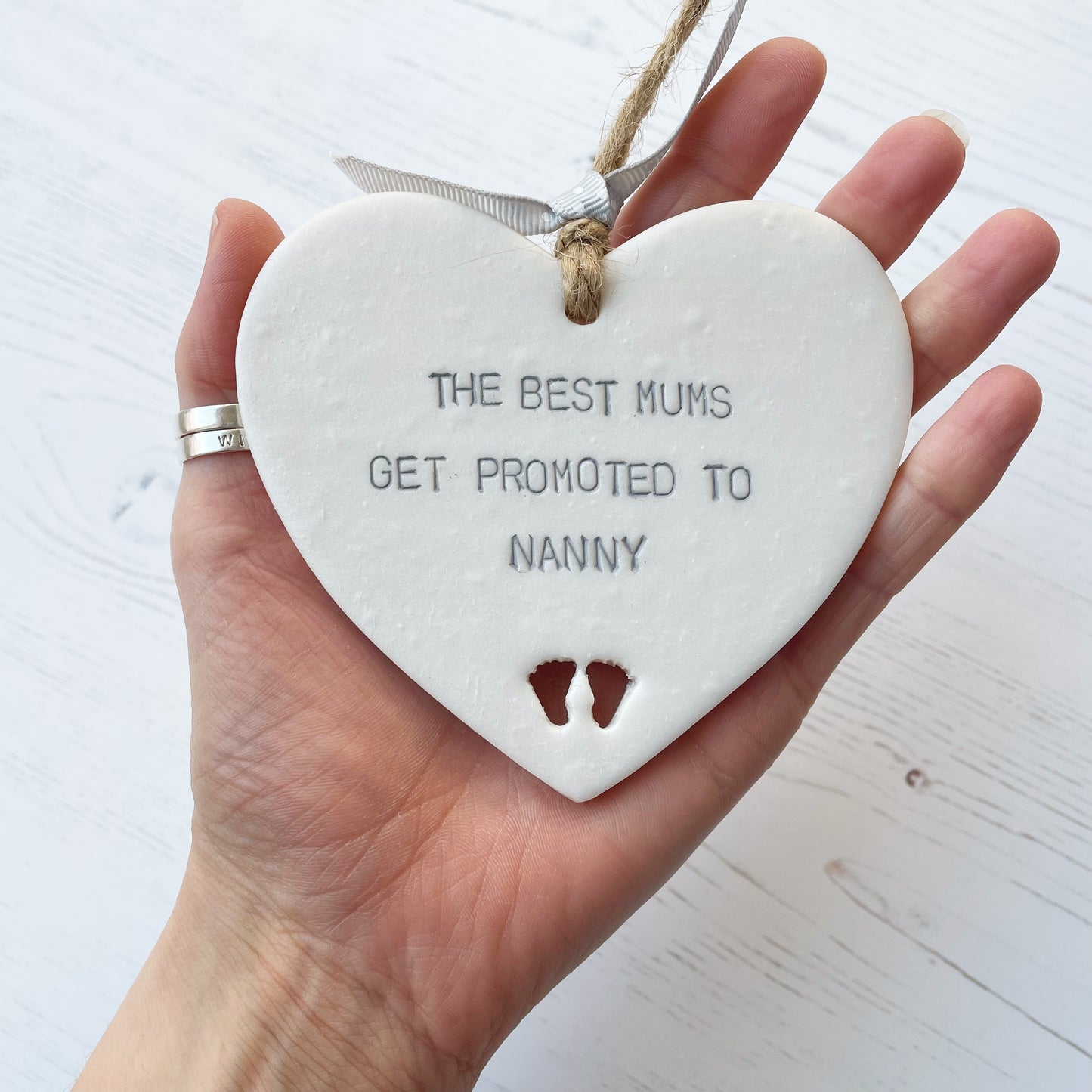 Pearlised white clay hanging heart with baby feet cut out of the bottom and grey personalisation, the heart is personalised with THE BEST MUMS GET PROMOTED TO NANNY