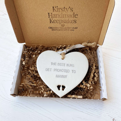 Pearlised white clay hanging heart with baby feet cut out of the bottom and grey personalisation, the heart is personalised with THE BEST MUMS GET PROMOTED TO NANNY
