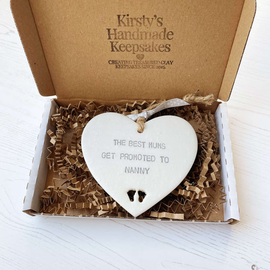 Pearlised white clay hanging heart with baby feet cut out of the bottom and grey personalisation, the heart is personalised with THE BEST MUMS GET PROMOTED TO NANNY