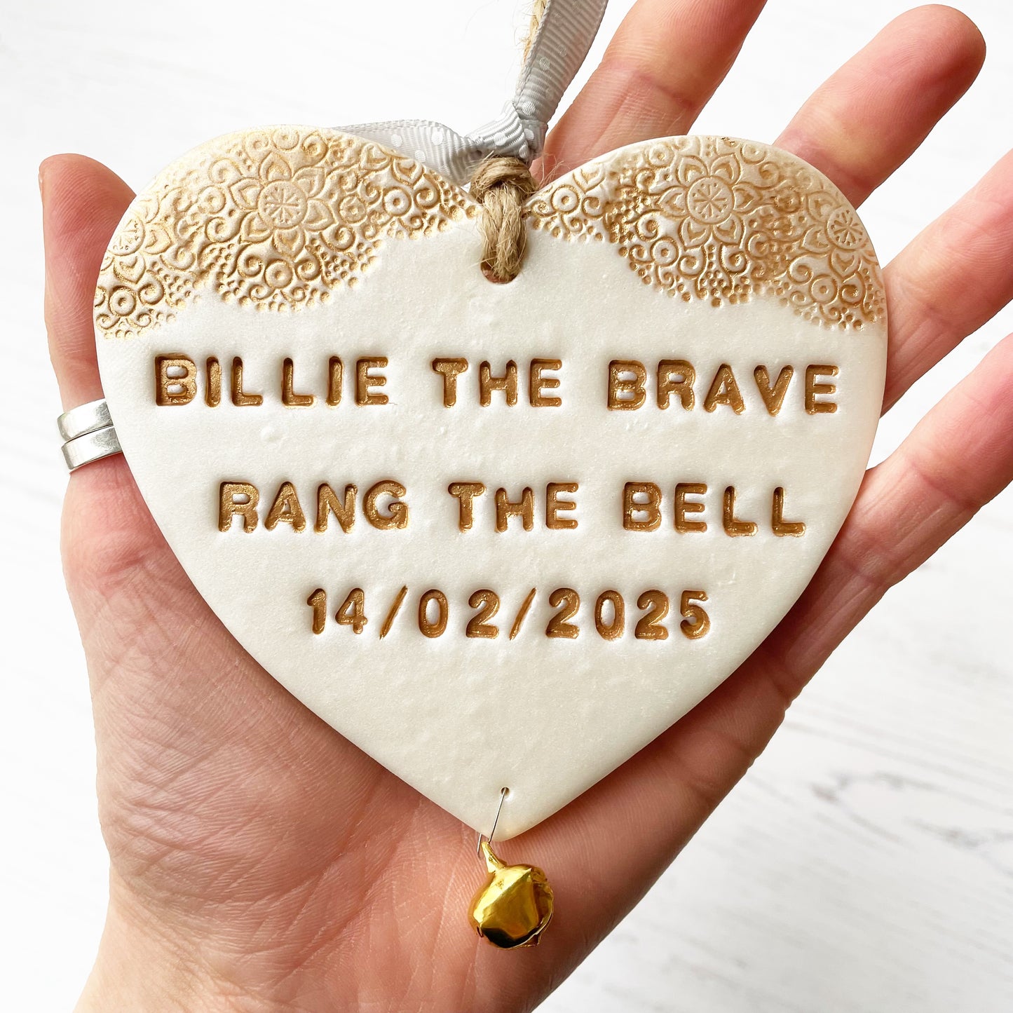 Personalised cancer survivor gift, pearlised white clay hanging heart with a gold lace edge at the top of the heart and a gold bell hanging below, the heart is personalised with BILLIE THE BRAVE RANG THE BELL 14/02/2025