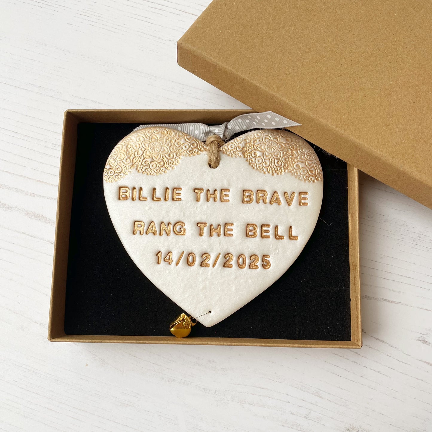 Personalised cancer survivor gift, pearlised white clay hanging heart with a gold lace edge at the top of the heart and a gold bell hanging below, the heart is personalised with BILLIE THE BRAVE RANG THE BELL 14/02/2025